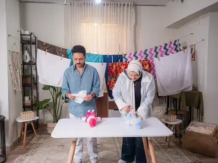 Two adults stand at a table changing diapers on baby dolls, with laundry hanging on a clothesline behind them in a home setting.