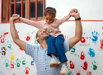 Father carries his daughter onto his shoulders while they play in front of a wall decorated with colourful handprints.