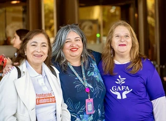 Three women pose together outside a building, with two wearing purple shirts featuring organization logos.