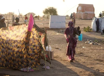 A woman holding a child, facing away from the camera, walks past makeshift tents