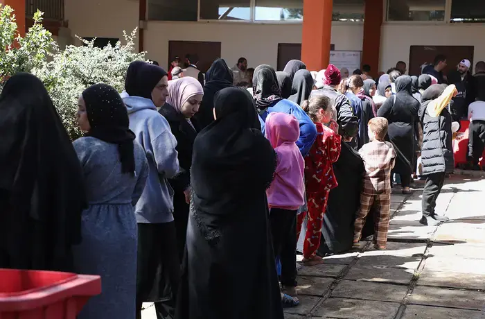 Women and children stand in a queue outdoors at a displacement shelter, waiting in line in a courtyard.