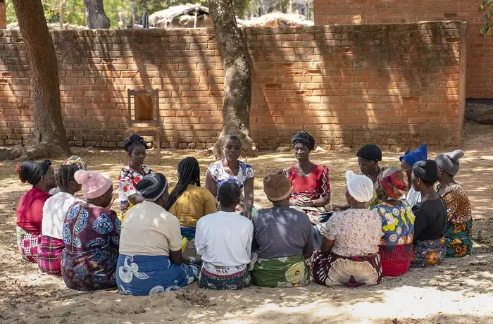 A group of women sit together on the ground, under the shade of a tree
