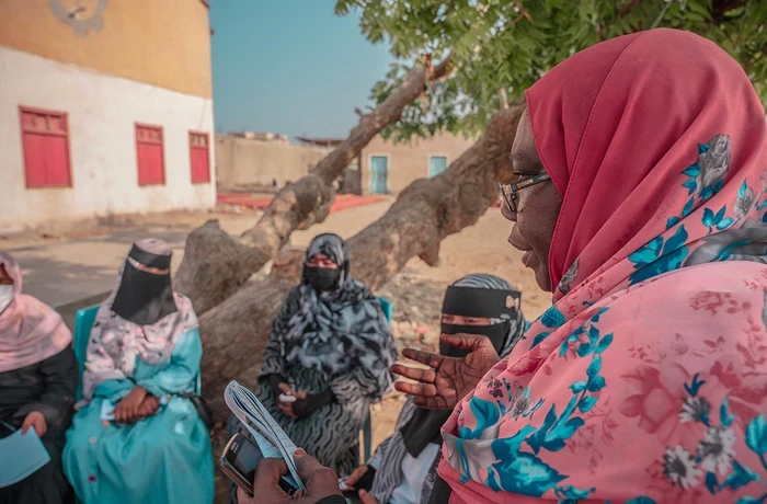 A woman speaks to a group of women seated outdoors, holding papers and a phone.