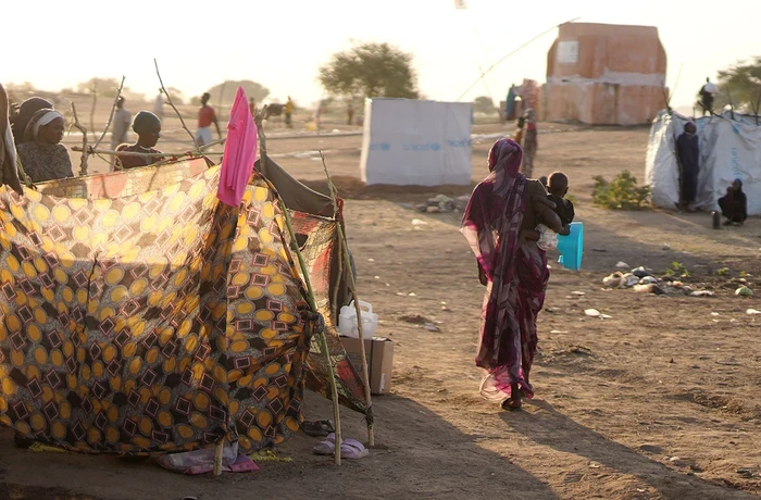A woman holding a child, facing away from the camera, walks past makeshift tents