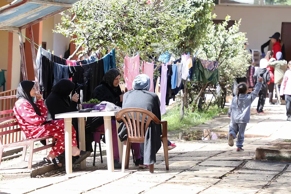 Women sit together outdoors near clotheslines while a child walks along a pathway in a displacement shelter.