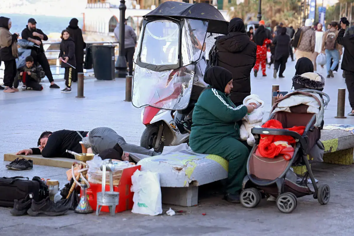 A woman sits beside a stroller caring for a child on a busy street, with a person lying nearby.