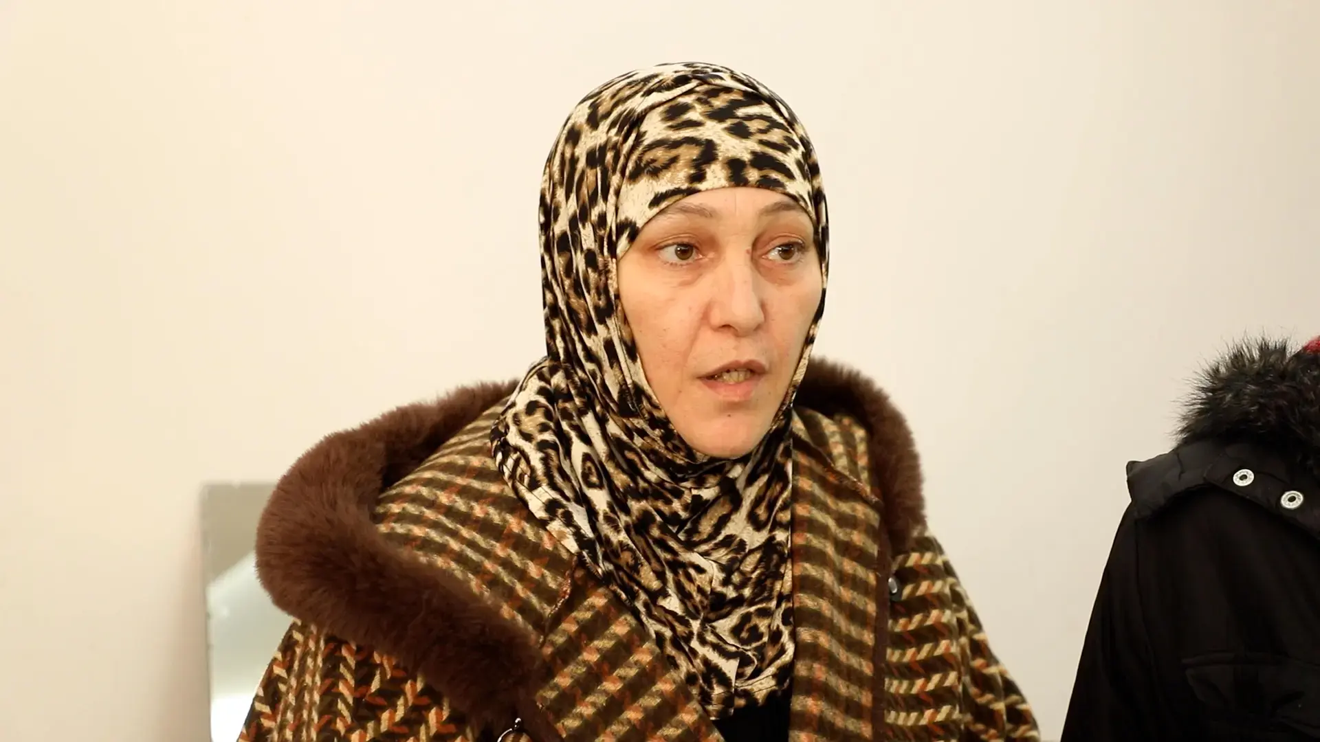 A woman speaks indoors while wearing a patterned headscarf and coat.