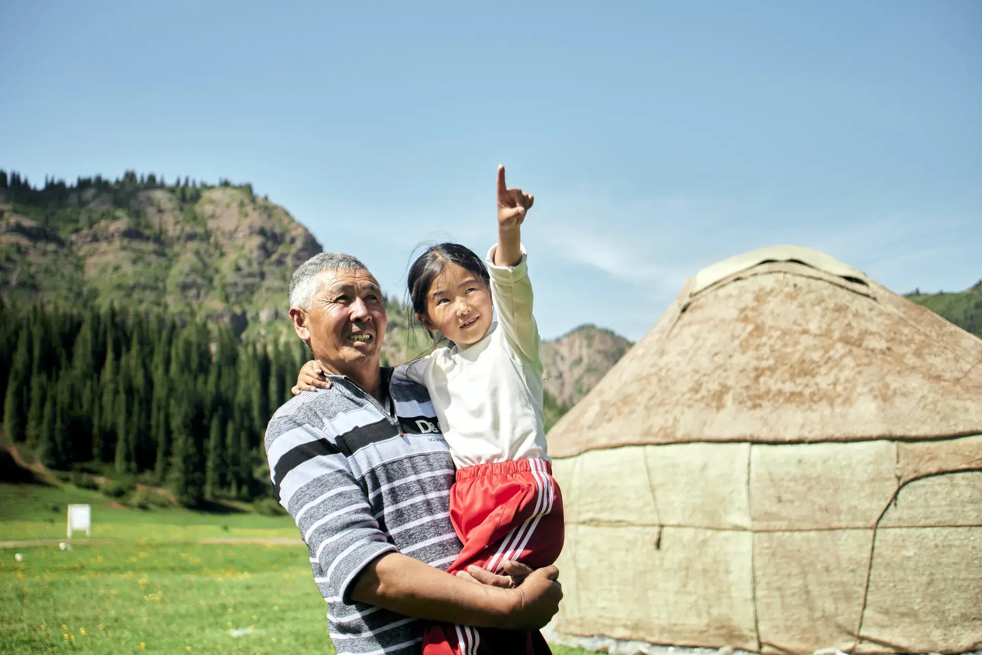 Un homme porte une petite fille qui pointe du doigt le ciel, alors qu'ils se trouvent dans un paysage verdoyant avec des montagnes et une tente en arrière-plan.