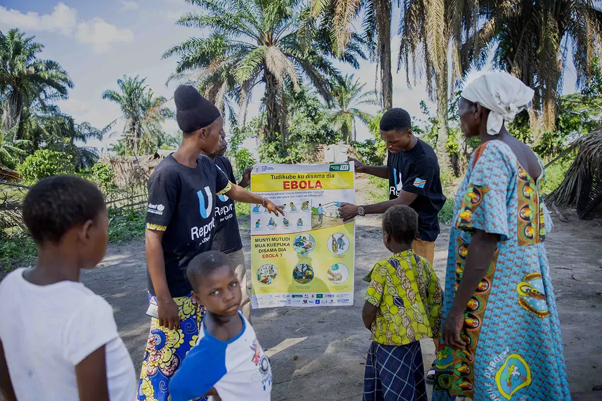 Deux personnes tiennent une affiche de sensibilisation au virus Ebola tout en discutant avec des membres de la communauté et des enfants rassemblés en plein air.