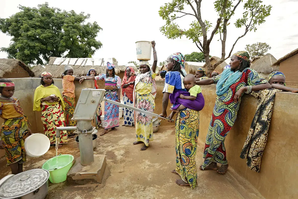 Des femmes et des enfants sont rassemblés autour d'une pompe à eau manuelle ; l'eau est recueillie dans des seaux et des récipients.