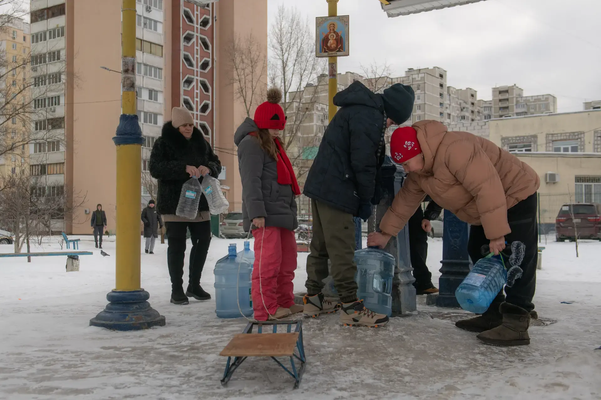Plusieurs personnes remplissent des récipients d'eau à un point d'eau public dans un quartier résidentiel enneigé.