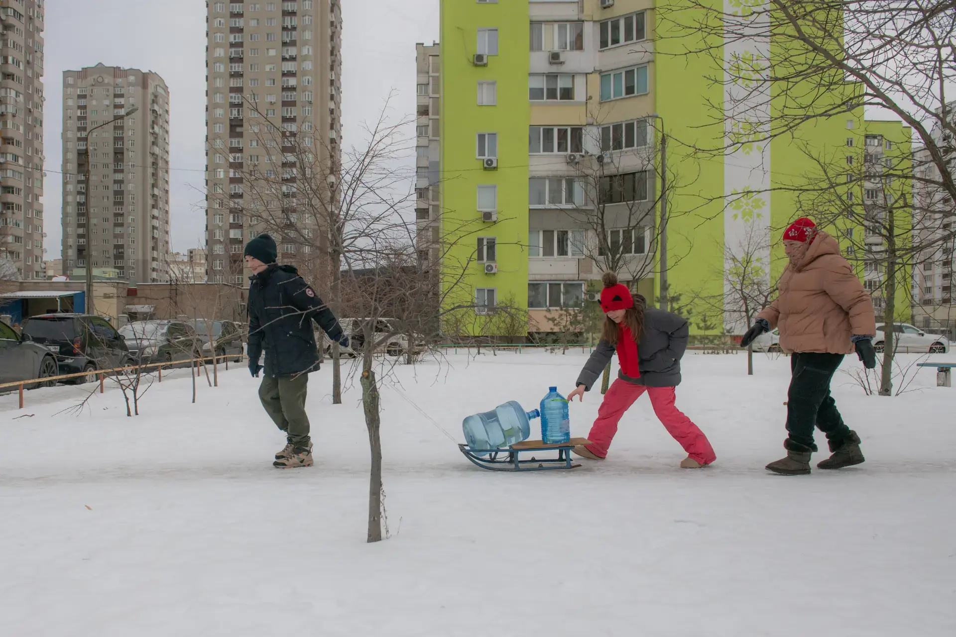 Trois personnes traversent un quartier résidentiel enneigé, tirant de grands bidons d'eau sur un traîneau entre deux immeubles.