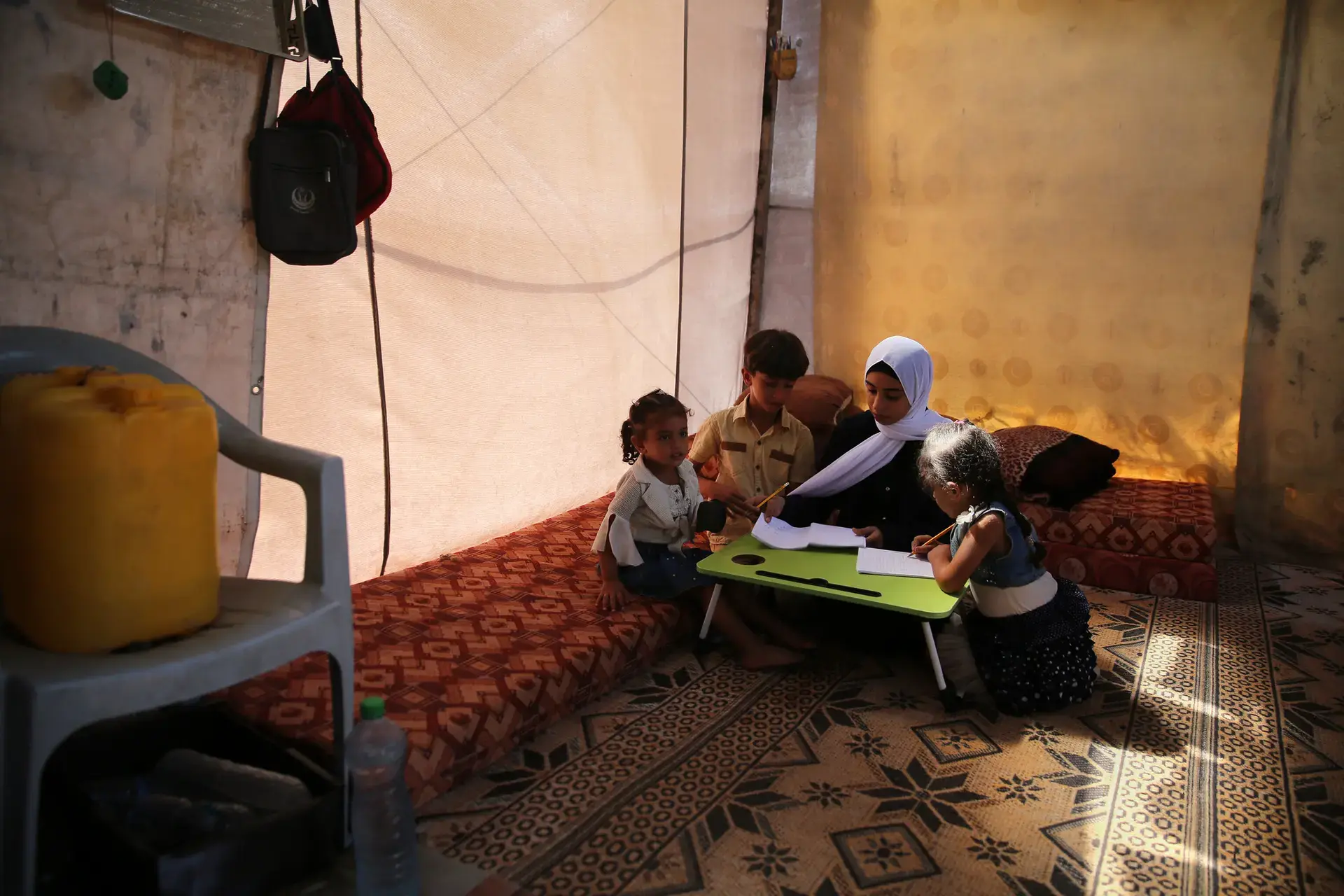 A girl tutors her three younger siblings around a small table inside a makeshift tent.