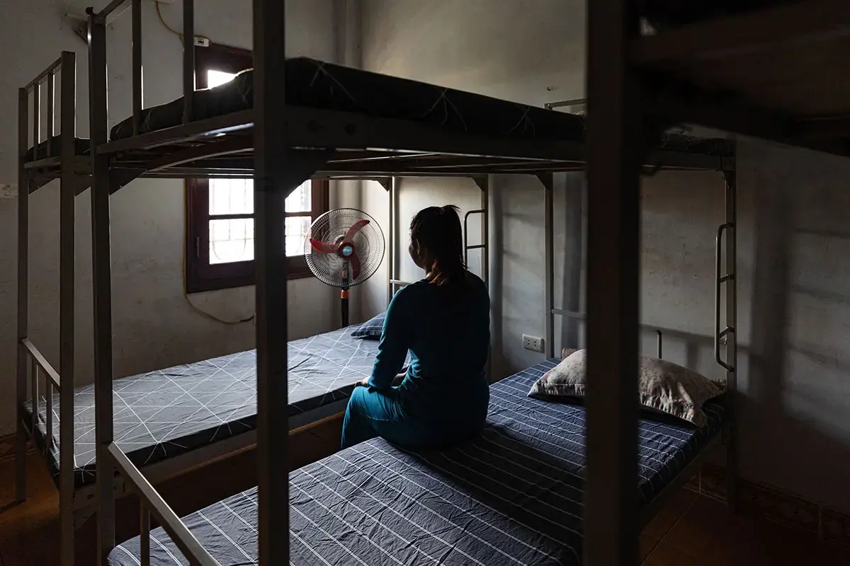 A woman sits alone on a bunk bed, facing a window with several empty bunk beds surrounding her.