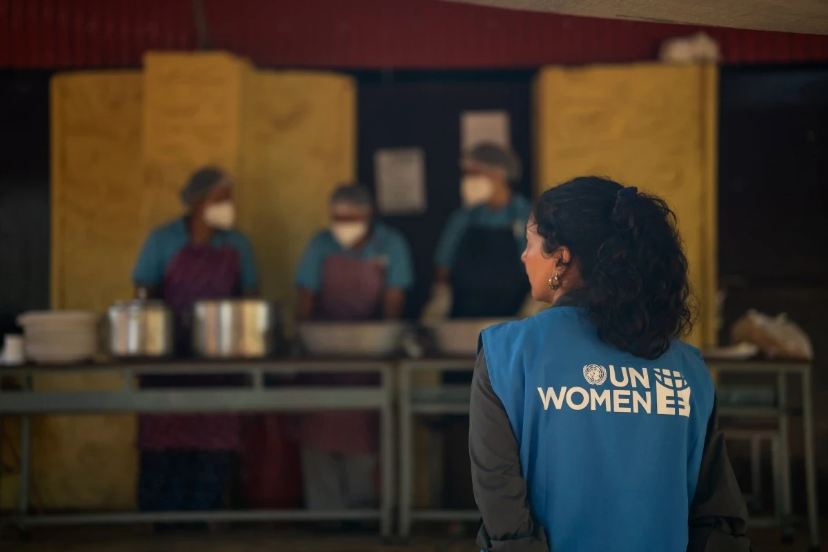 A woman wearing a blue UN Women vest stands with chefs as they prepare meals