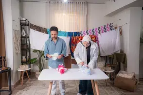 Two adults stand at a table changing diapers on baby dolls, with laundry hanging on a clothesline behind them in a home setting.